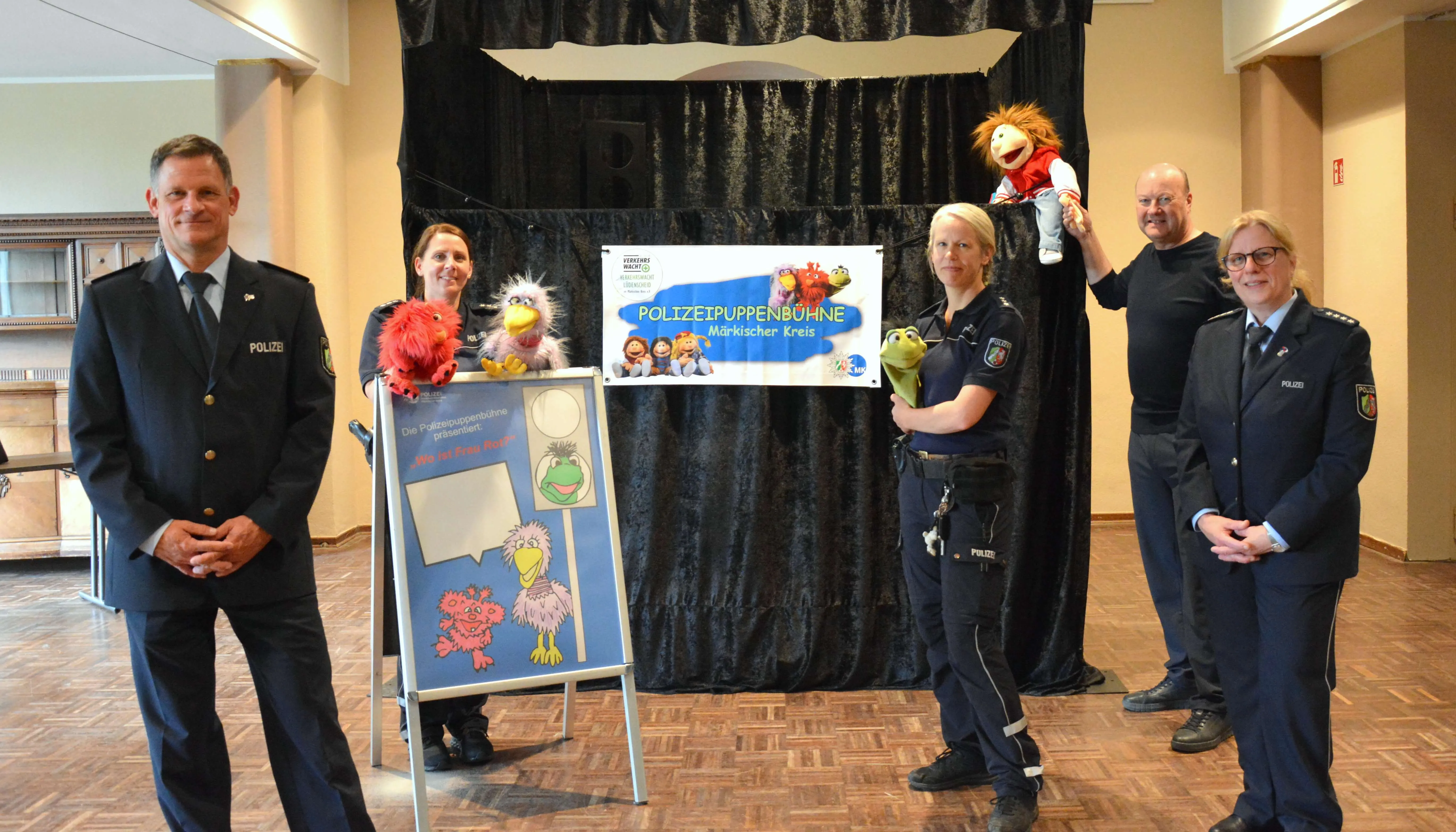 Fünf Polizeibeamte stehen vor einem Banner mit der Aufschrift „Polizeipuppenbühne Märkischer Kreis“ und halten Handpuppen. Im Vordergrund ein Schild mit kindlichen Illustrationen zu Verkehrssicherheit.