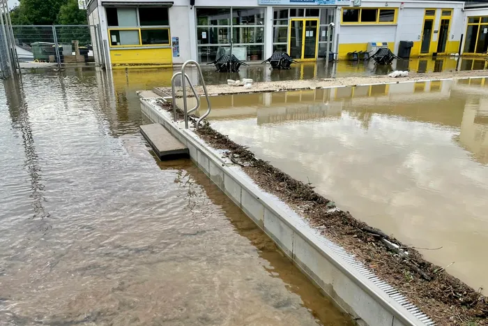 Überflutetes Schwimmbecken vor einem Gebäude mit gelber Fassade. Die Wasseroberfläche spiegelt den Himmel wider.