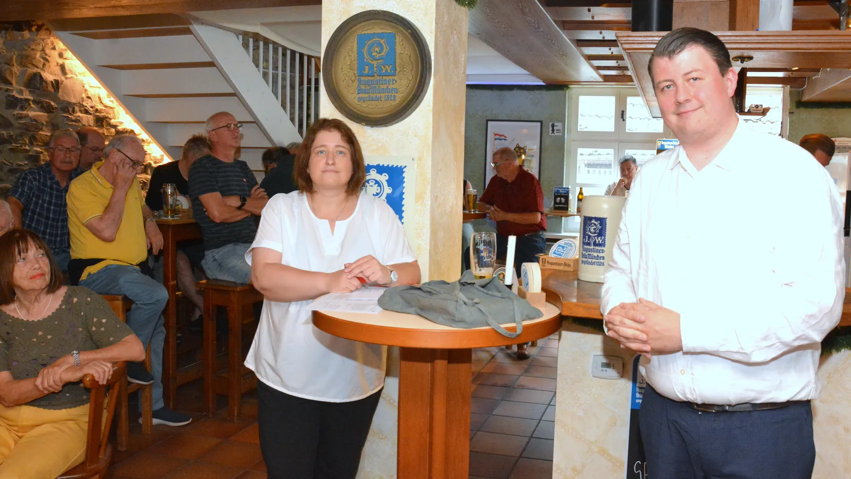 Zwei Personen, eine Frau und ein Mann, stehen im Restaurant „Kater August“ und sprechen. Im Hintergrund sitzen weitere Gäste an Tischen und an der Bar.