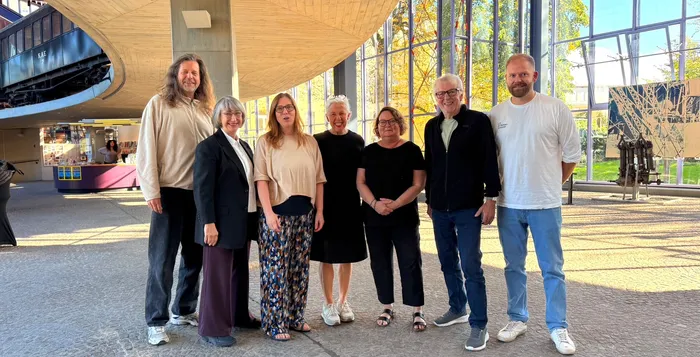 Die sieben Mitglieder der Jury stehen nebeneinander in einem hellen, modernen Gebäude mit großen Glasfronten.