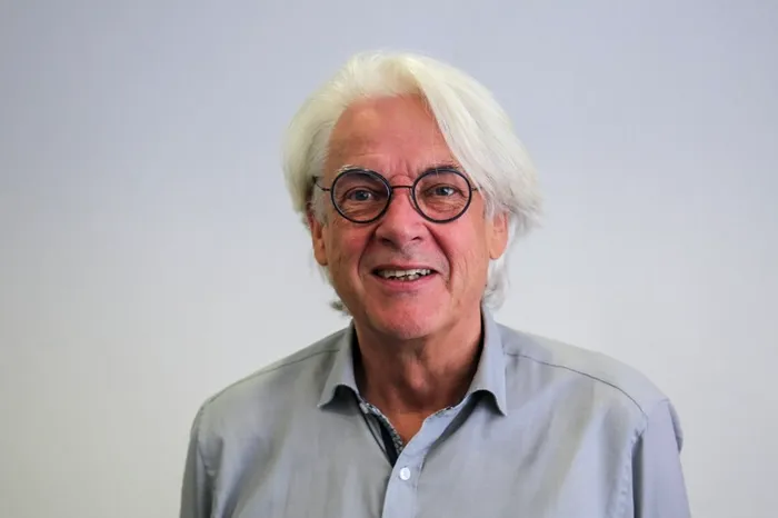 Rainer Danne mit weißem Haar und Brille lächelt vor einem hellen Hintergrund.
