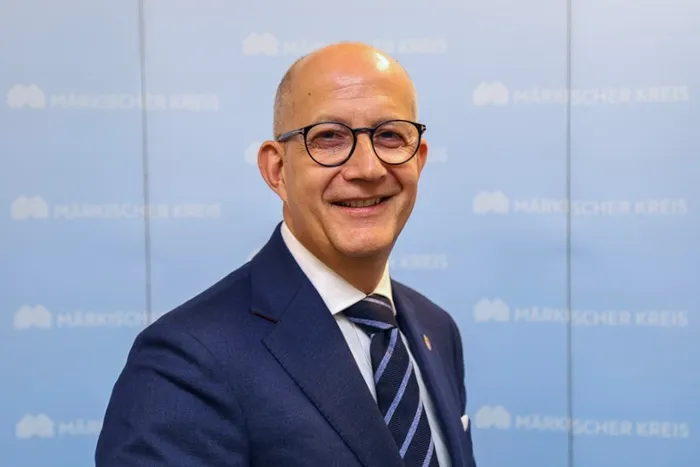 Ralf Schwarzkopf mit Brille und dunklem Anzug lächelt vor einem hellblauen Hintergrund mit dem Schriftzug „Märkischer Kreis“.