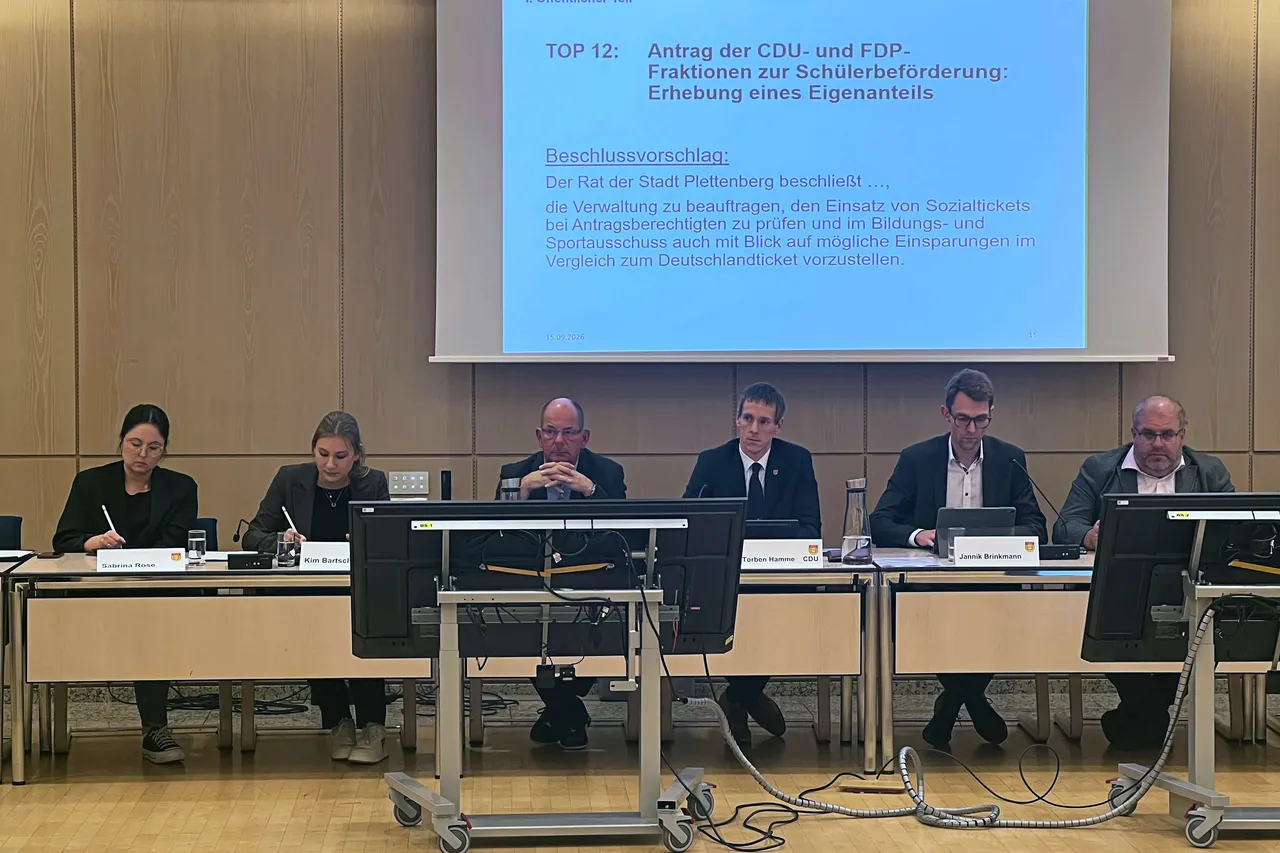Sechs Personen sitzen an einem Tisch in einem Ratssaal vor einer Projektionsleinwand mit einer Präsentation.