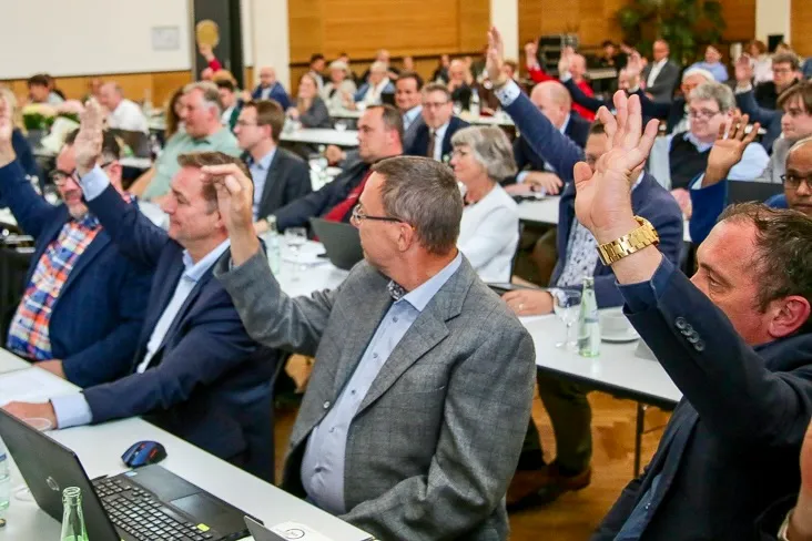 Versammlung von Ratsmitgliedern bei einer Abstimmung, viele heben die Hand. Im Vordergrund ein Mann mit Brille und blauem Hemd.
