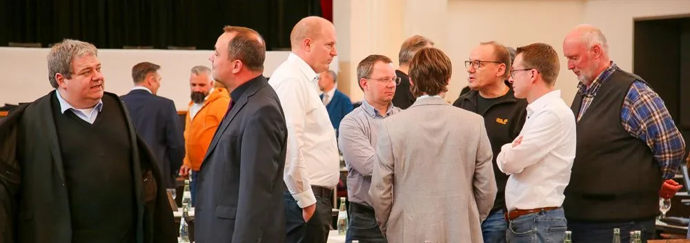 Eine Gruppe von Männern steht in einem Raum zusammen und unterhält sich. Auf Tischen sind Wasserflaschen zu sehen.