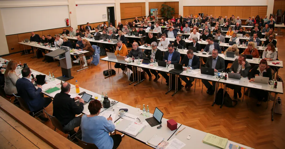 Großer Konferenzraum mit zahlreichen Teilnehmern an Tischen, die in einer U-Form angeordnet sind. Einige Personen stehen und sprechen, andere sitzen und arbeiten an Dokumenten oder Laptops.