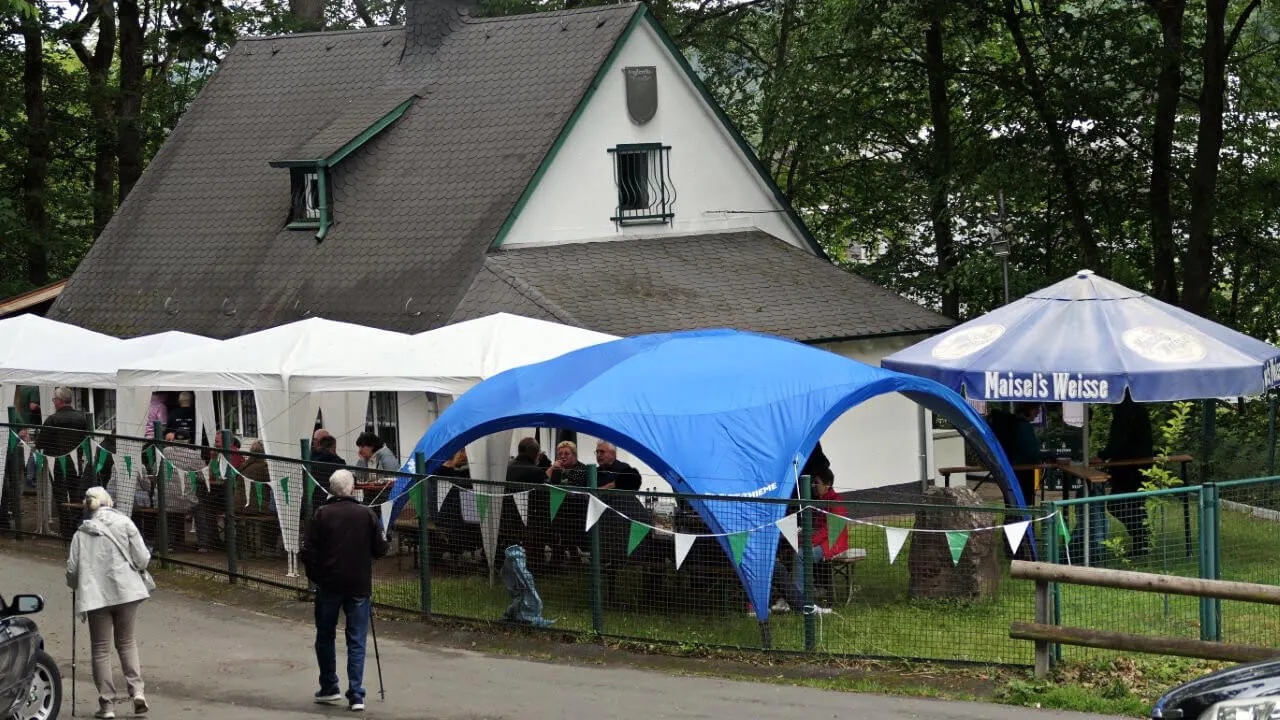 Menschen sitzen unter Pavillons und Sonnenschirmen vor einem Fachwerkhaus, das Räucherfest des Anglerclubs Forelle Eiringhausen.