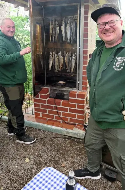 Zwei Männer in grünen Jacken stehen vor einem Räucherofen, in dem Fische hängen. Im Hintergrund ein Schild mit einem Spruch über das Angeln.