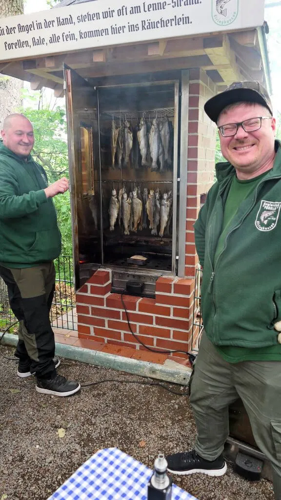 Zwei Männer in grünen Jacken stehen vor einem Räucherofen, in dem Fische hängen. Im Hintergrund ein Schild mit einem Spruch über das Angeln.