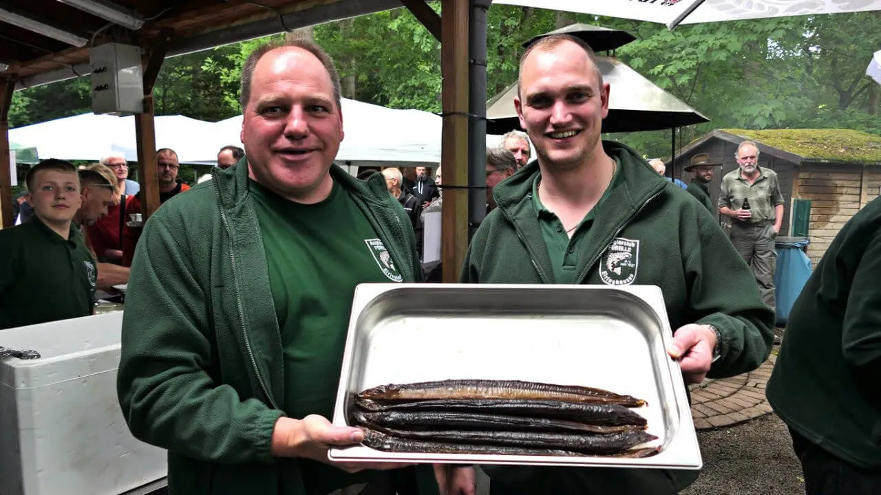 Zwei Männer in grünen Jacken halten eine silberne Platte mit geräucherten Forellen im Freien. Im Hintergrund sind weitere Personen und ein Festzelt zu sehen.