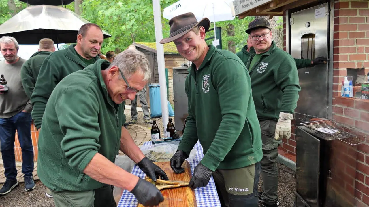 Mehrere Männer in grünen Jacken bereiten auf einem Holztisch vor einem Grill Fisch zu. Im Hintergrund steht ein Gebäude und weitere Personen sind zu sehen.