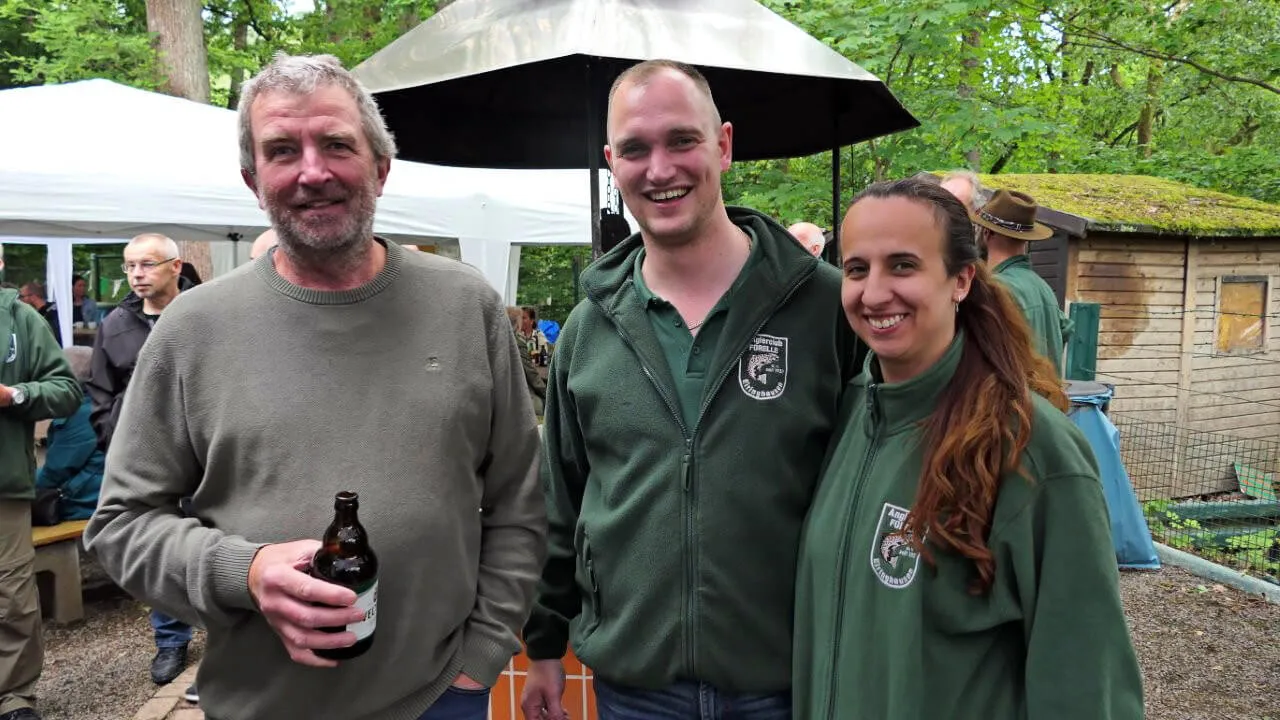 Drei lächelnde Menschen stehen vor einem Schirm und einem Gartenhaus. Der Mann links hält eine Bierflasche, die beiden anderen tragen grüne Jacken mit einem Fisch-Logo.