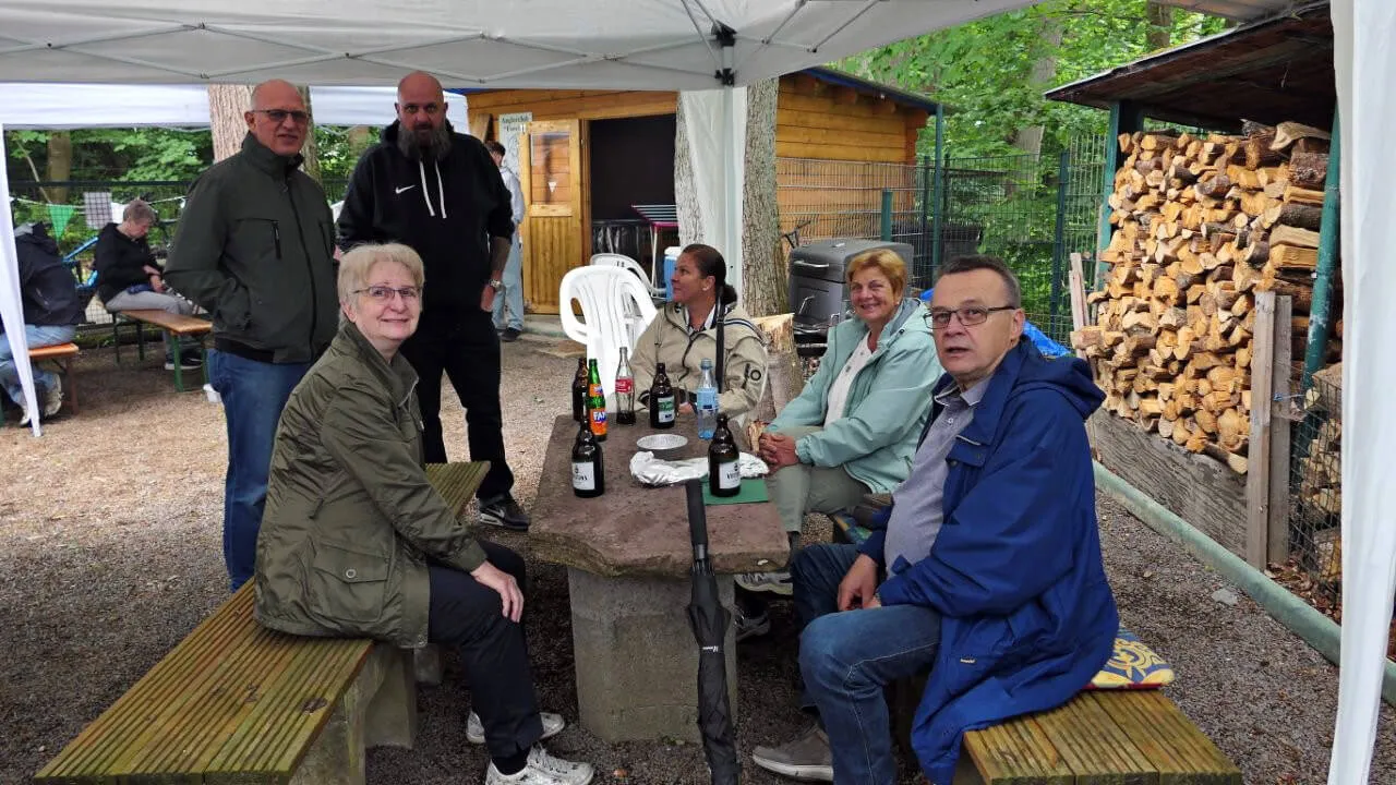 Mehrere Personen sitzen an einem Holztisch im Freien, umgeben von Bierflaschen und Holzhaufen. Im Hintergrund ein Zelt und weitere Personen.
