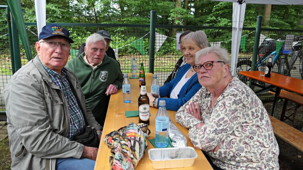 Vier ältere Menschen sitzen an einem Biergartentisch unter einem weißen Zelt und unterhalten sich. Auf dem Tisch stehen Getränke und eine Snackbox.