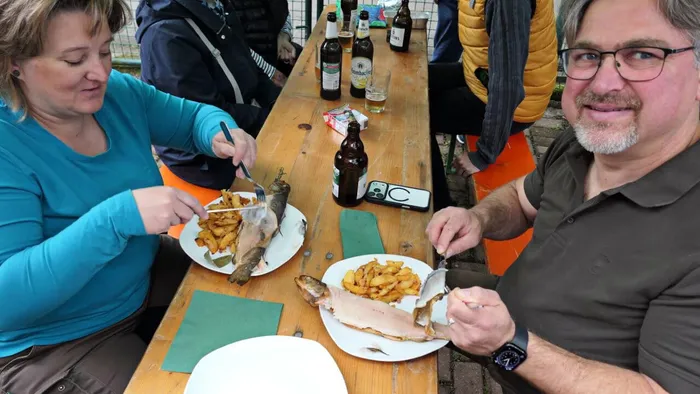 Zwei Personen essen gebratene Forelle mit Bratkartoffeln an einem langen Holztisch im Freien, umgeben von Bierflaschen und weiteren Personen.