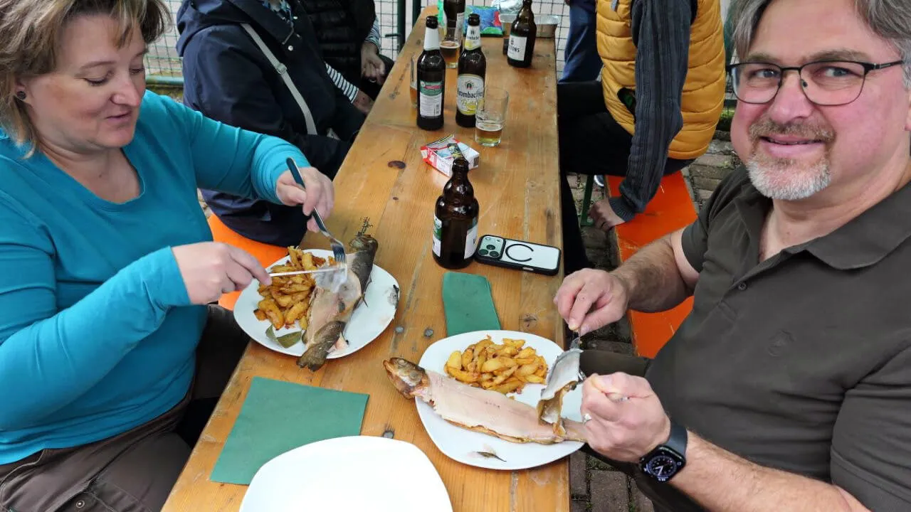 Zwei Personen essen gebratene Forelle mit Bratkartoffeln an einem langen Holztisch im Freien, umgeben von Bierflaschen und weiteren Personen.