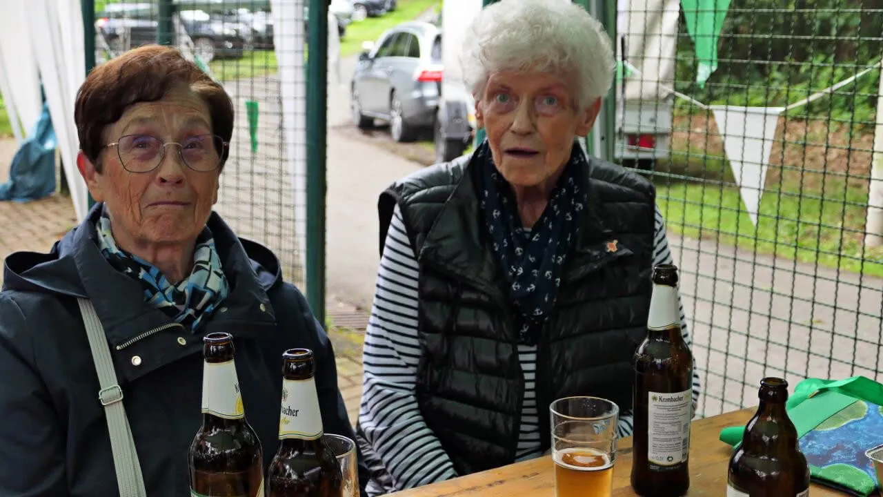 Zwei ältere Frauen sitzen an einem Tisch mit Bierflaschen und einem gefüllten Glas. Im Hintergrund ein Tennisplatz und weitere Personen.