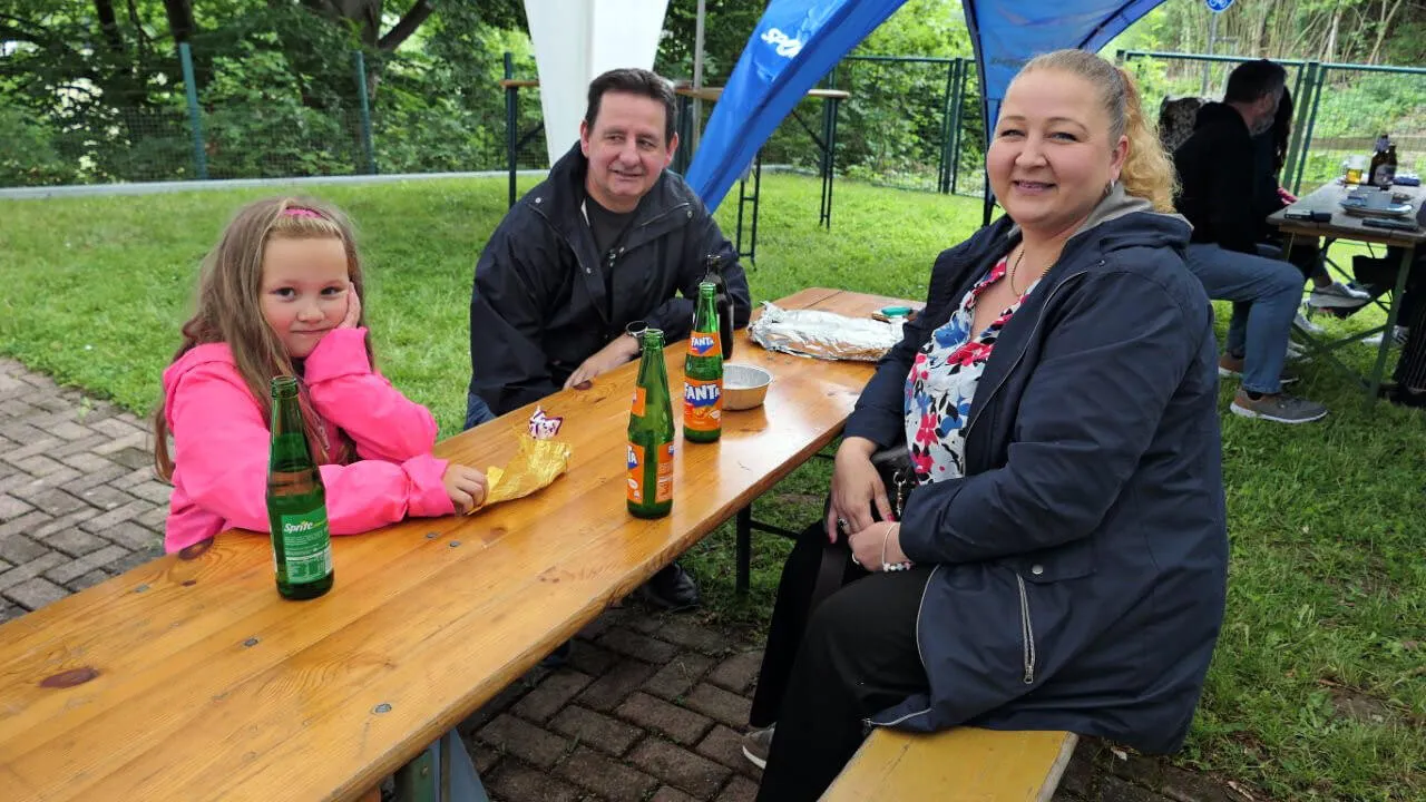 Drei Personen sitzen an einem Holztisch im Freien, vor ihnen stehen Getränkeflaschen. Im Hintergrund sind weitere Personen und ein blaues Zelt zu sehen.