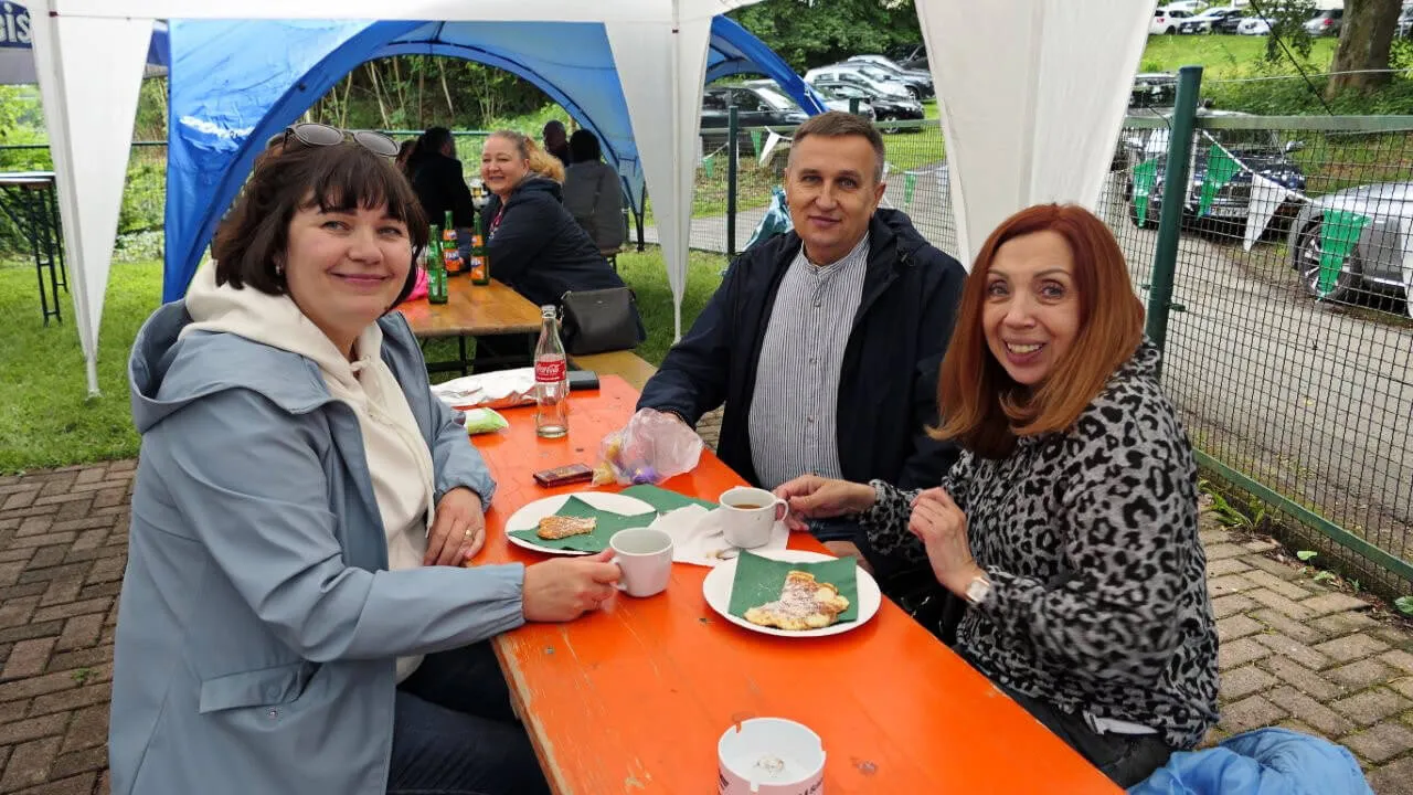 Drei Personen sitzen an einem orangefarbenen Tisch im Freien und essen von Tellern mit Speisen. Im Hintergrund sind Zelte und weitere Personen zu sehen.