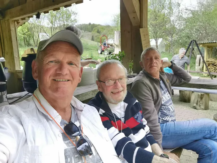 Drei lächelnde Männer sitzen im Freien unter einem Holzpavillon. Im Hintergrund ist ein Traktor auf einer Wiese zu sehen.