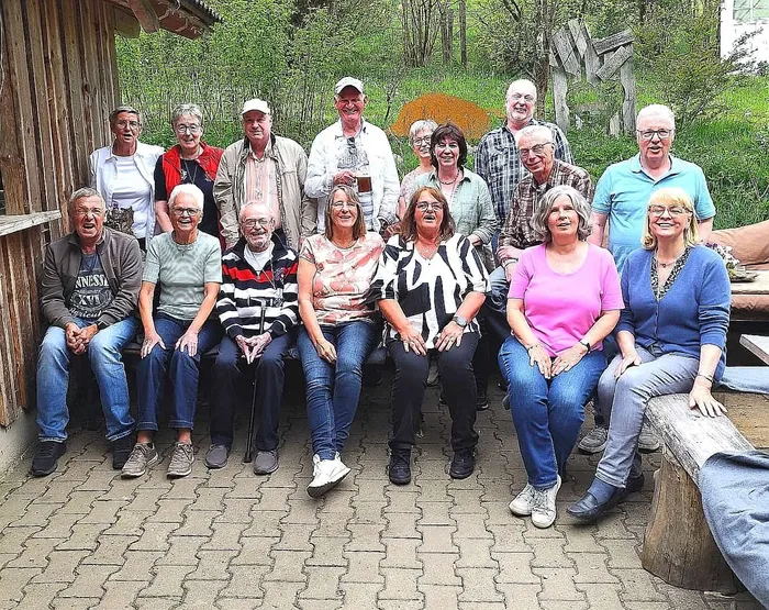 Eine Gruppe von etwa 15 ehemaligen Schülern und ihrem Klassenlehrer posiert lächelnd für ein Gruppenfoto im Freien.