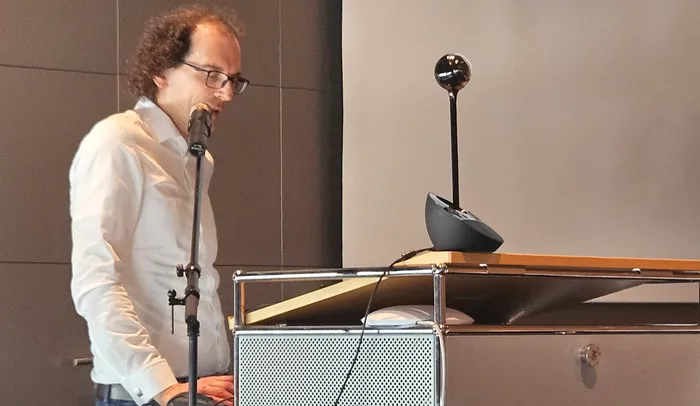 Ein Mann mit lockigem Haar und Brille spricht in ein Mikrofon an einem Pult. Im Hintergrund eine graue Wand.