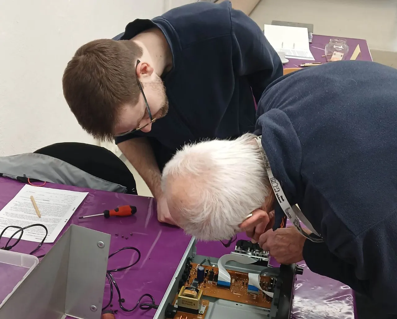 Zwei Personen beugen sich über ein geöffnetes elektronisches Gerät und reparieren es mit Werkzeugen.