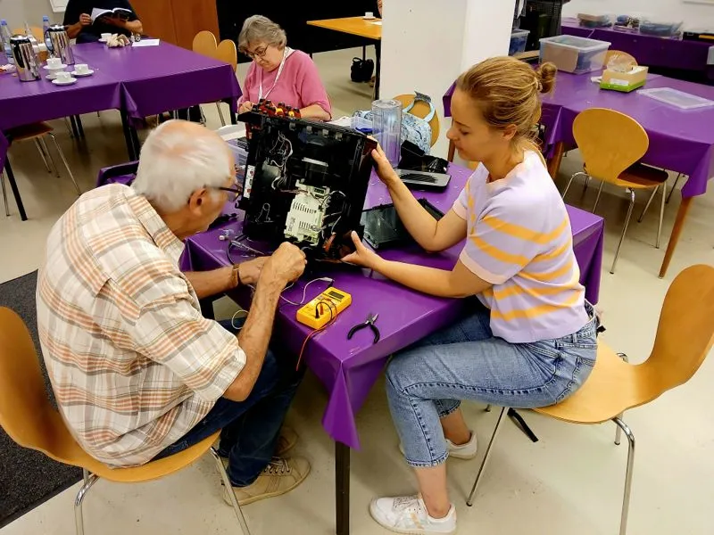 Ein älterer Mann und eine junge Frau reparieren gemeinsam ein elektronisches Gerät an einem Tisch mit lila Tischdecke. Im Hintergrund weitere Personen an Tischen.