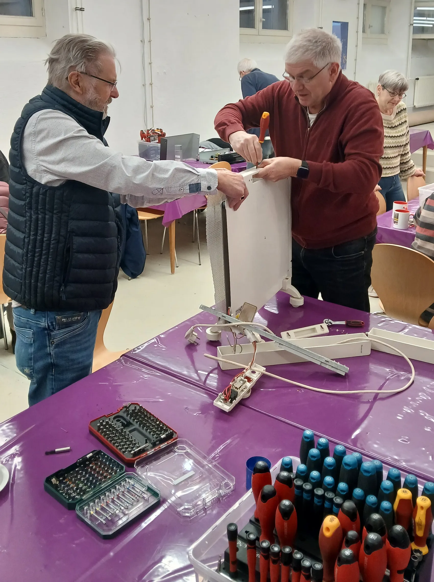 Zwei Männer reparieren gemeinsam ein elektrisches Gerät an einem violett bedeckten Tisch, umgeben von Werkzeug und weiteren Helfern.