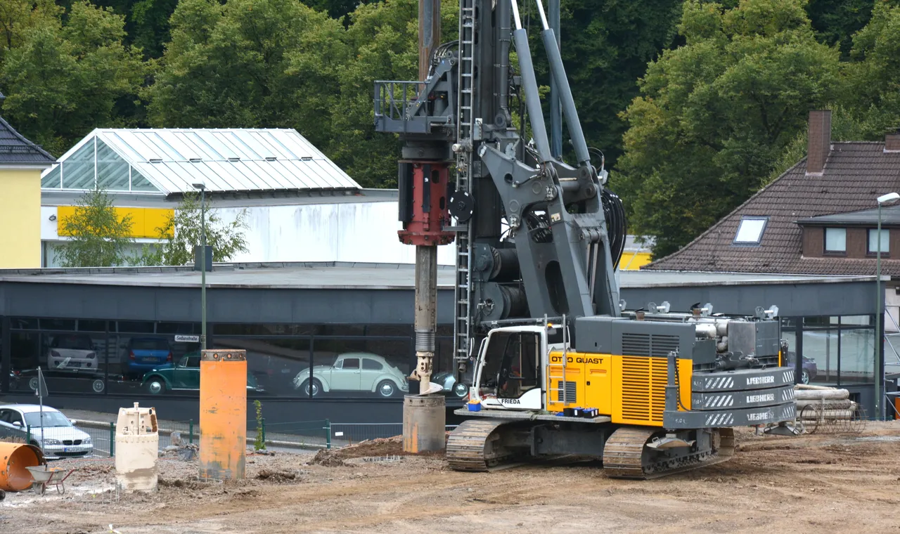 Ein großer grauer Bohrer steht auf einer Baustelle vor einem Gebäude mit einem hellen Dach und einem weißen Kleinwagen davor. Im Hintergrund sind weitere Gebäude und Bäume zu sehen.