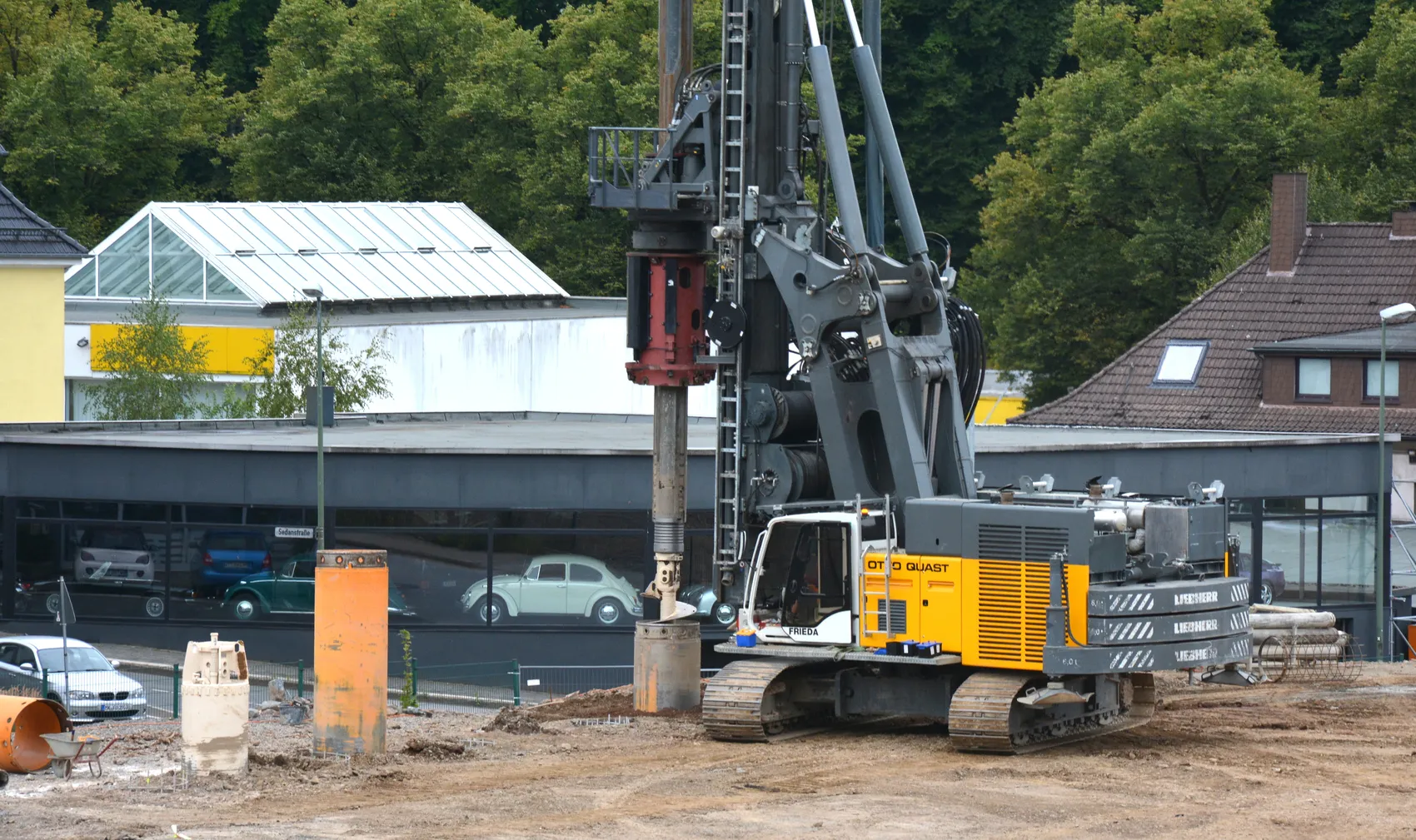 Ein großer grauer Bohrer steht auf einer Baustelle vor einem Gebäude mit einem hellen Dach und einem weißen Kleinwagen davor. Im Hintergrund sind weitere Gebäude und Bäume zu sehen.