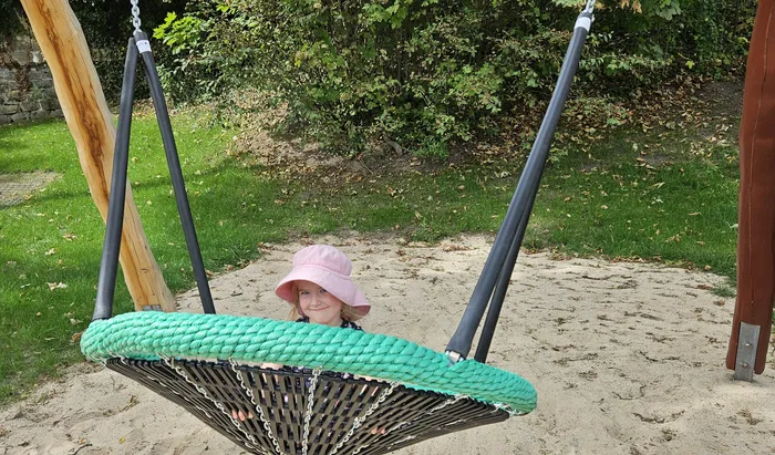 Ein Kind mit rosa Hut sitzt in einer grünen Nestschaukel auf einem Sandplatz.