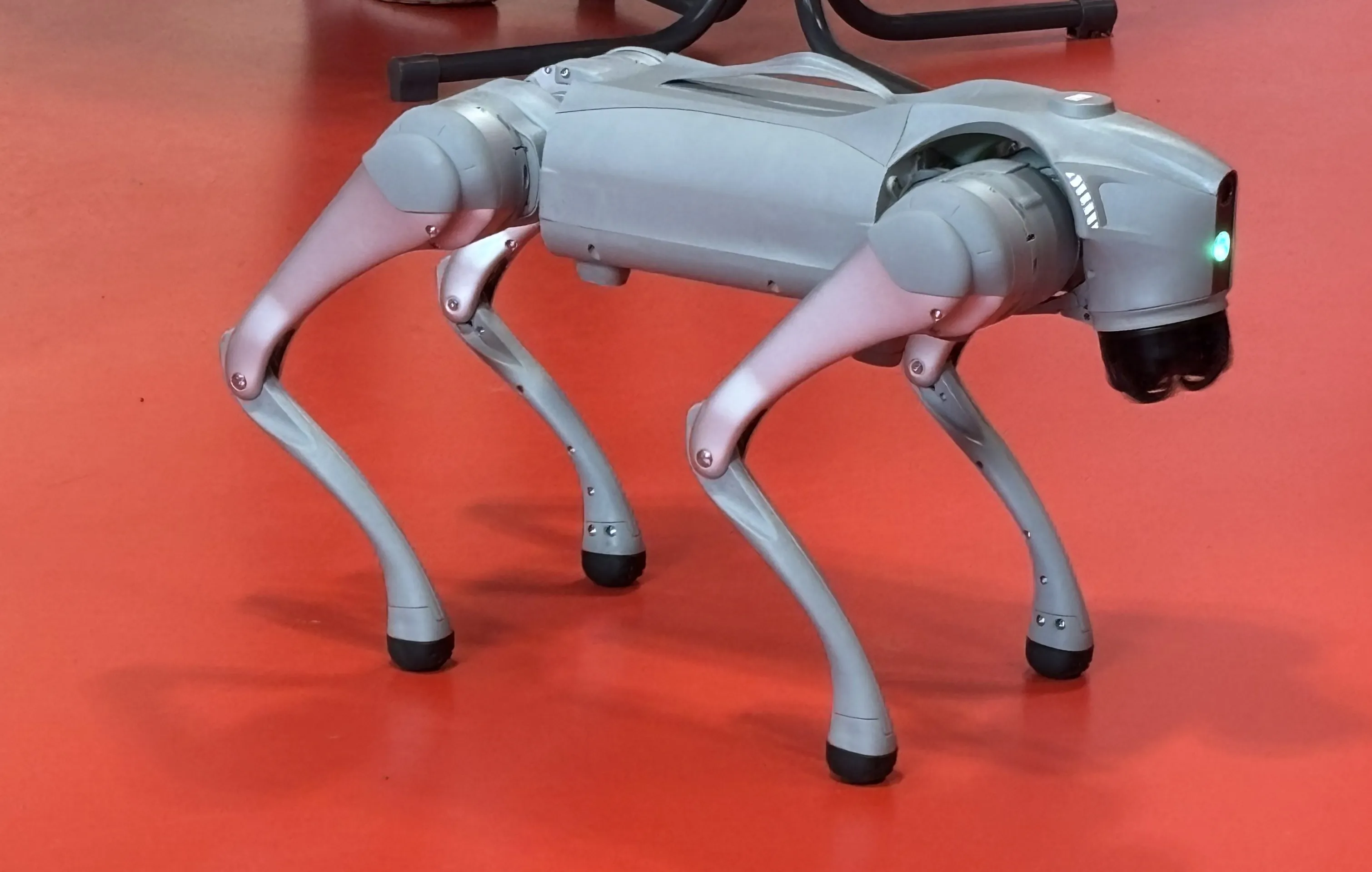 Silberner Roboterhund steht auf orangefarbenem Boden und blickt mit einer Kamera nach vorne. Das Gerät hat vier Beine und eine komplexe mechanische Struktur.