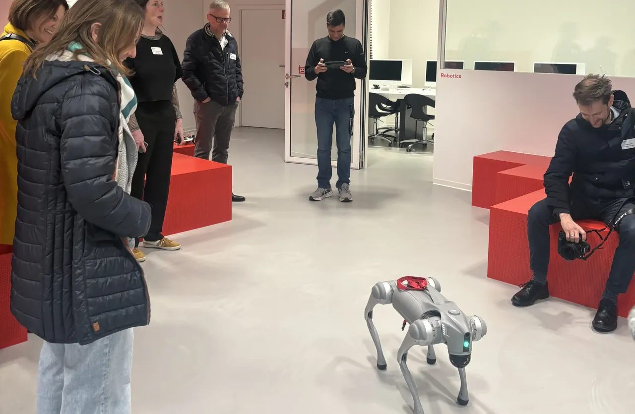Mehrere Personen beobachten einen vierbeinigen Roboter in einem hellen Raum. Im Hintergrund ist ein Arbeitsbereich mit Computern und dem Schriftzug „Robotics“ zu sehen.