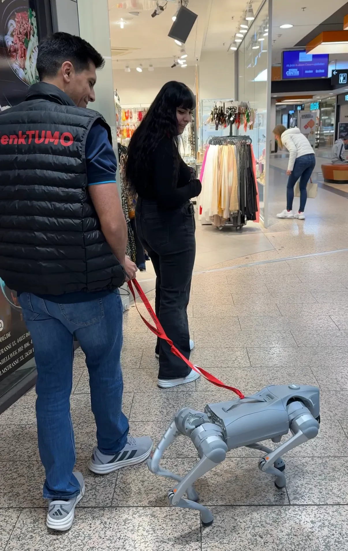 Ein Mann und eine Frau beobachten einen vierbeinigen Roboter, der an einer Leine geführt wird. Im Hintergrund sind Geschäfte und Passanten in einer Einkaufsstraße zu sehen.