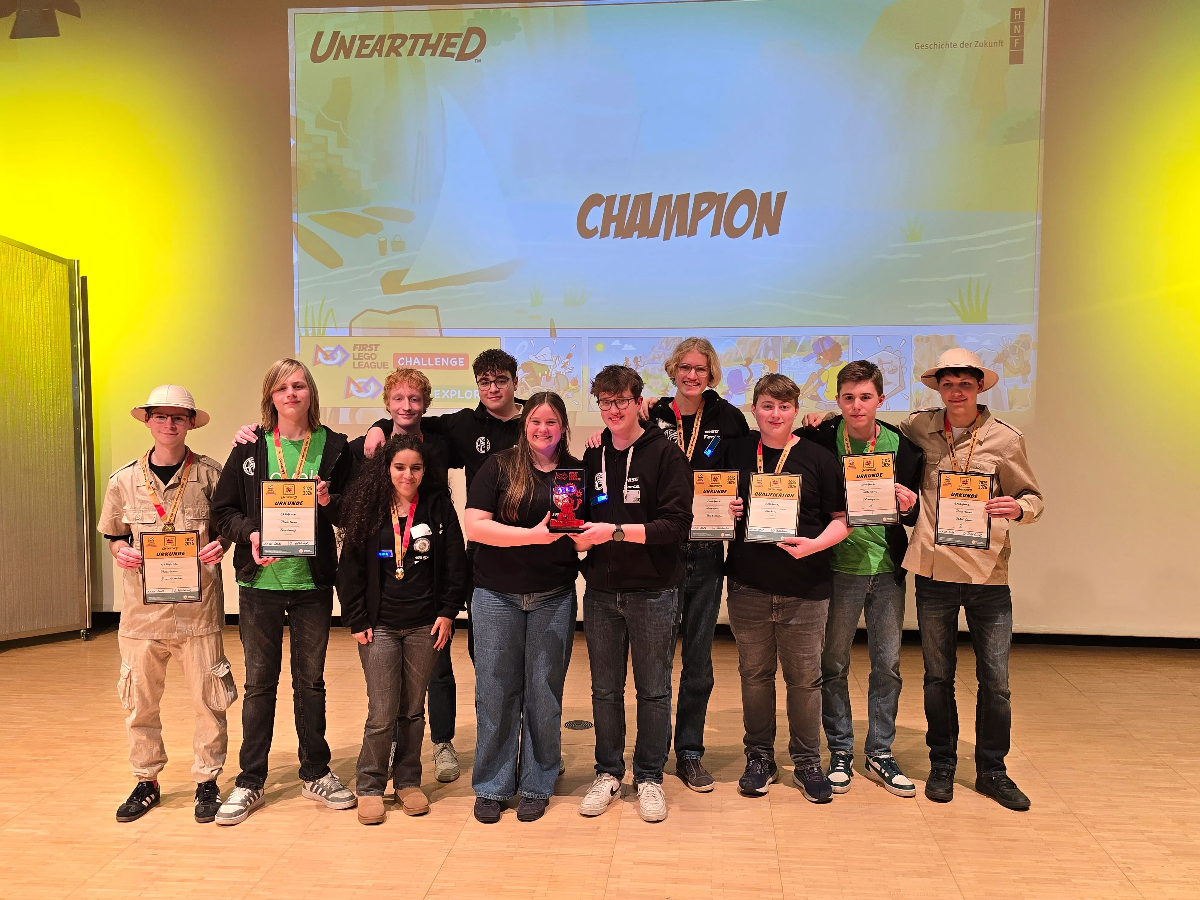 Eine Gruppe von zehn Jugendlichen steht auf einer Bühne und hält Urkunden mit der Aufschrift „1. Platz“ in der Hand. Im Hintergrund ist eine Leinwand mit dem Schriftzug „Champion“ und einer Illustration einer Bohrinsel zu sehen.