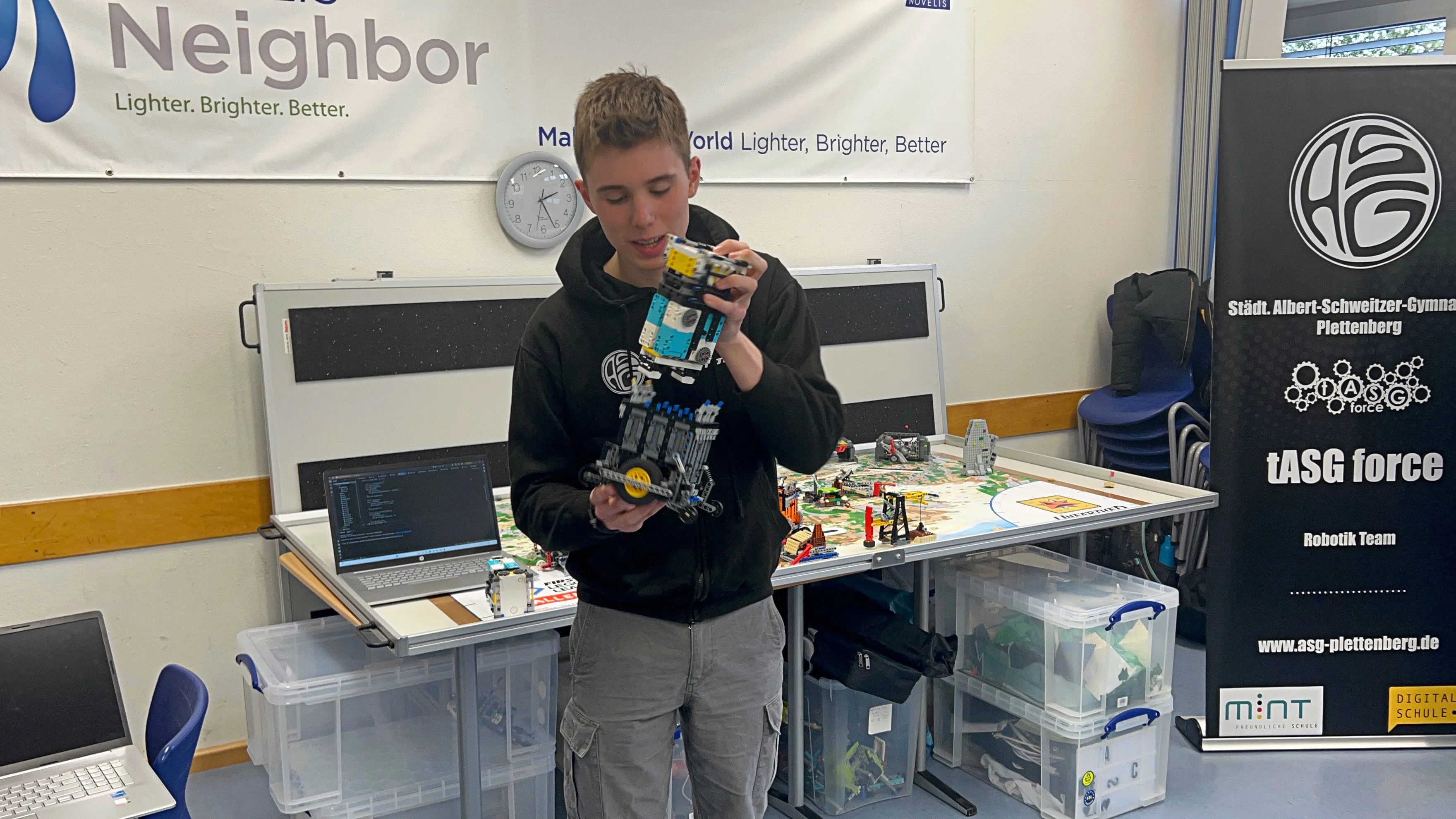 Ein Jugendlicher hält einen komplexen Lego-Roboter in der Hand, während er an einem Laptop vor einer Tischkonstruktion mit weiteren Lego-Elementen arbeitet. Im Hintergrund ein Banner mit dem Logo der Robotik-AG tASG force des Albert-Schweitzer-Gymnasiums Plettenberg.