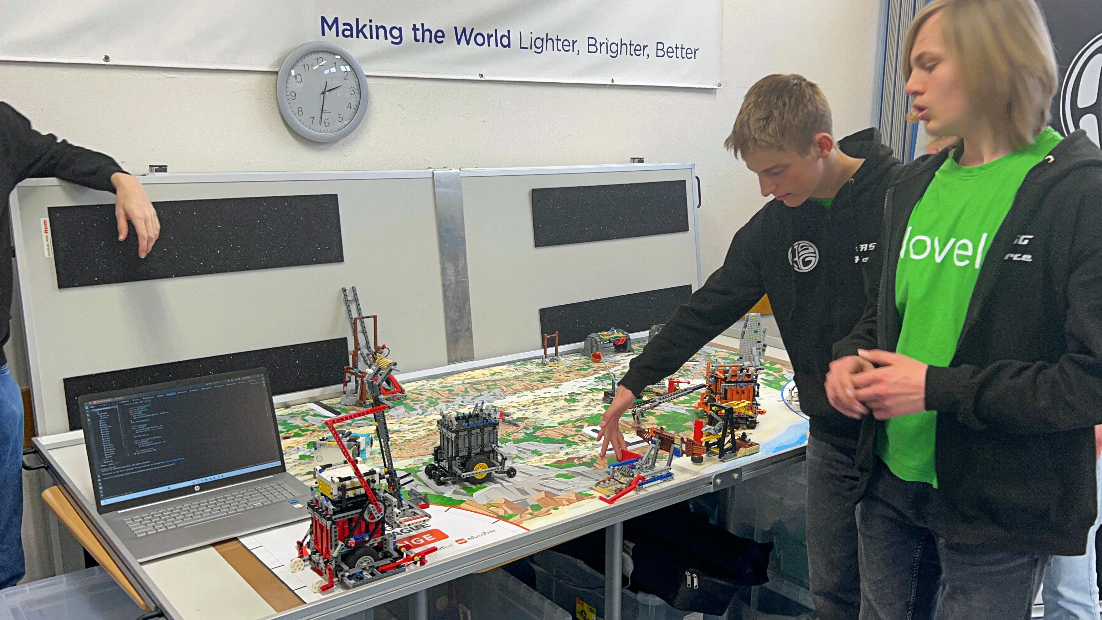 Drei Jugendliche arbeiten an einem Lego-Roboter auf einem Tisch mit einer Stadtlandschaft aus Lego-Steinen. Im Hintergrund ein Laptop mit Code und ein Spruch: „Making the World Lighter, Brighter, Better“.