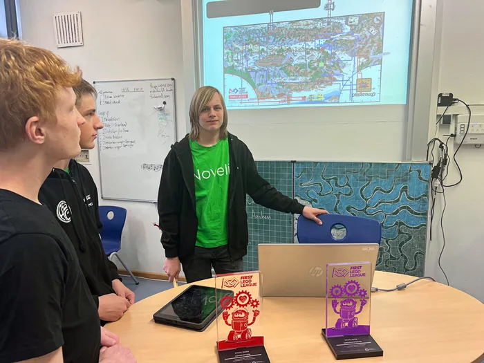 Drei Jugendliche stehen in einem Klassenzimmer vor einer Projektion einer Karte und besprechen etwas. Im Vordergrund stehen Auszeichnungen der First Lego League.