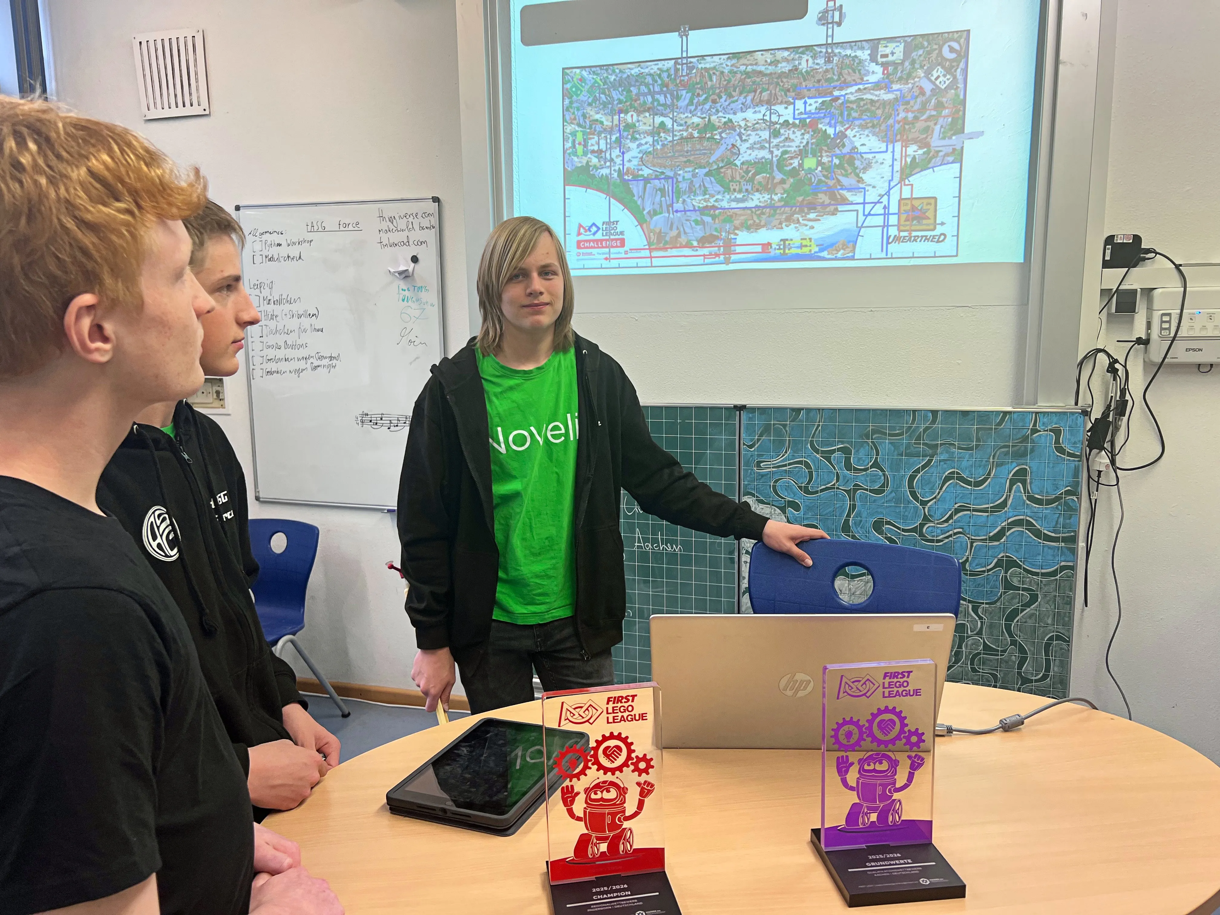 Drei Jugendliche stehen in einem Klassenzimmer vor einer Projektion einer Karte und besprechen etwas. Im Vordergrund stehen Auszeichnungen der First Lego League.