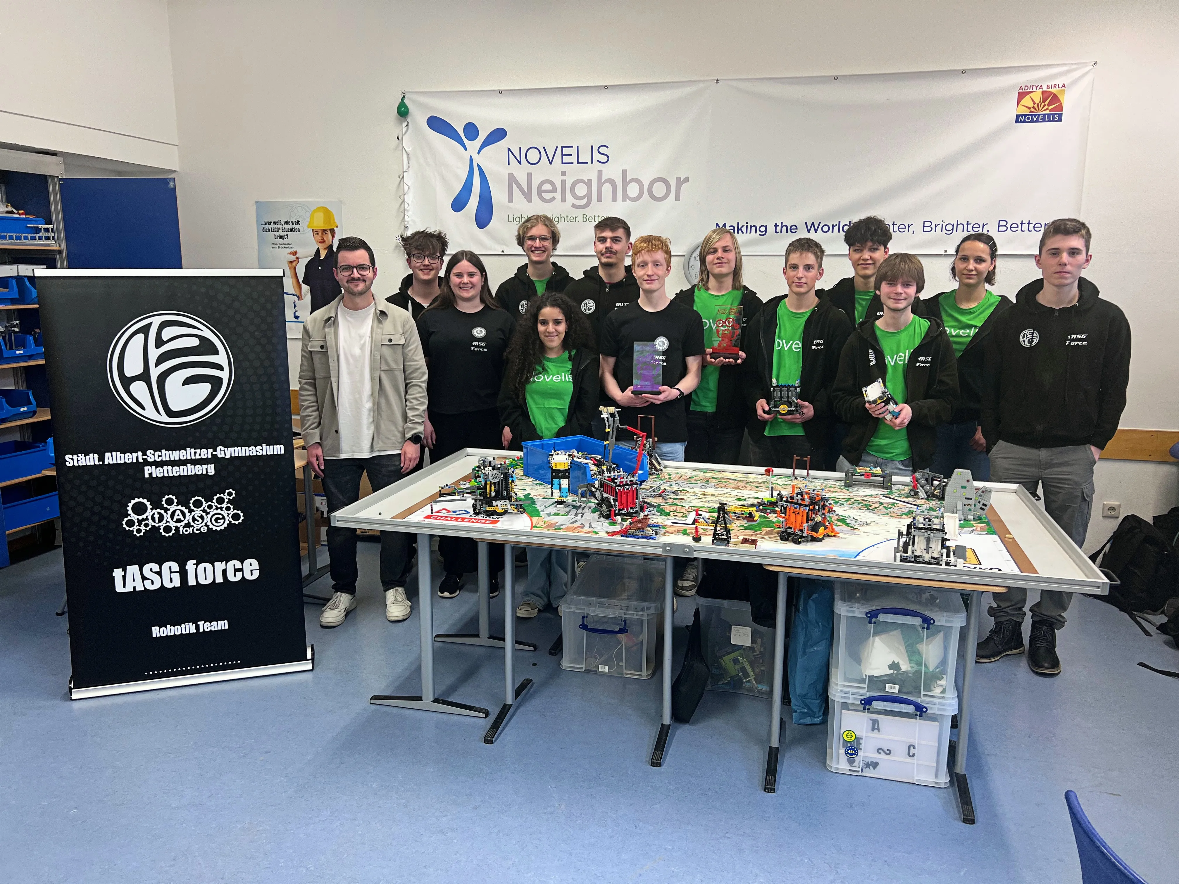 Eine Gruppe von Schülerinnen und Schülern steht vor einem Tisch mit einem aufgebauten Lego-Roboter. Im Hintergrund ein Banner mit dem Logo von Novelis und dem Albert-Schweitzer-Gymnasium Plettenberg.