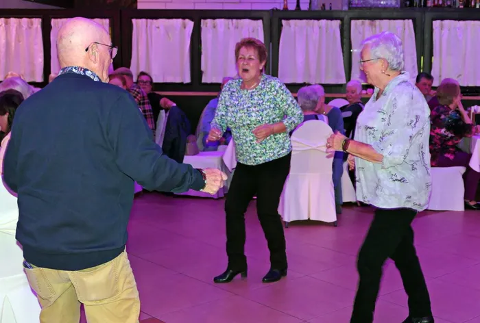 Ältere Menschen tanzen ausgelassen bei einer Party in einem Saal mit weißer Tischwäsche.