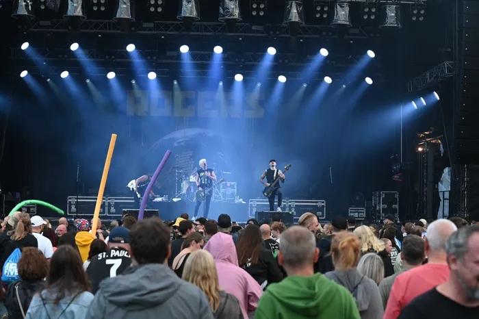 Die Band Rogers spielt unter blauen Scheinwerfern auf einer beleuchteten Bühne vor einer Menschenmenge.