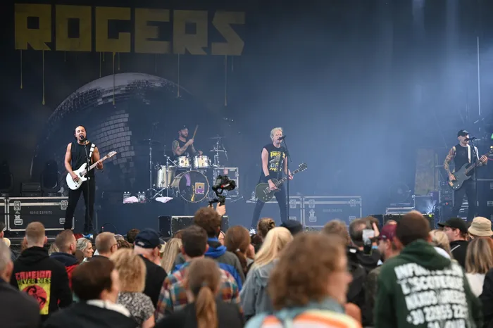 Die Band Rogers tritt live auf einer Bühne vor einem Publikum auf, im Hintergrund sind der Schriftzug „ROGERS“ und eine große Discokugel zu sehen.