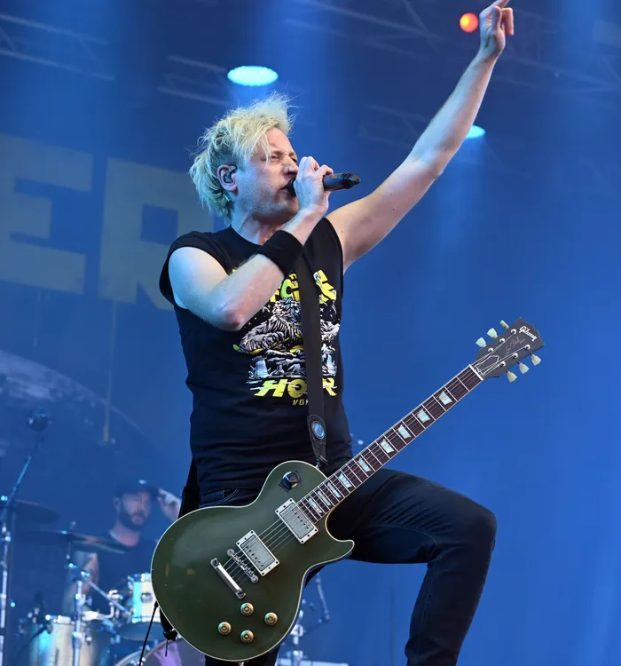 Rogers singt mit erhobenem Arm in ein Mikrofon auf einer Bühne vor einer dunkelgrünen E-Gitarre.