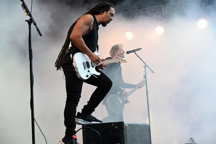 Ein Musiker mit Dreadlocks spielt eine weiße E-Gitarre auf einer vernebelten Bühne, während im Hintergrund ein weiterer Musiker singt.