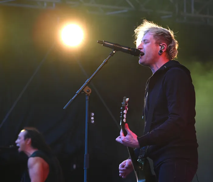 Ein Sänger mit blonden Haaren singt in ein Mikrofon und spielt Gitarre auf einer hell beleuchteten Bühne.