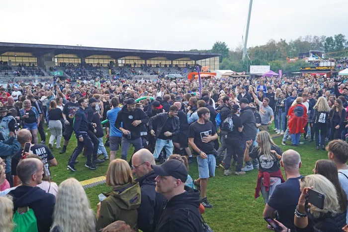 Eine große Menschenmenge auf einer Wiese vor einer überdachten Bühne bei einem Musikfestival.