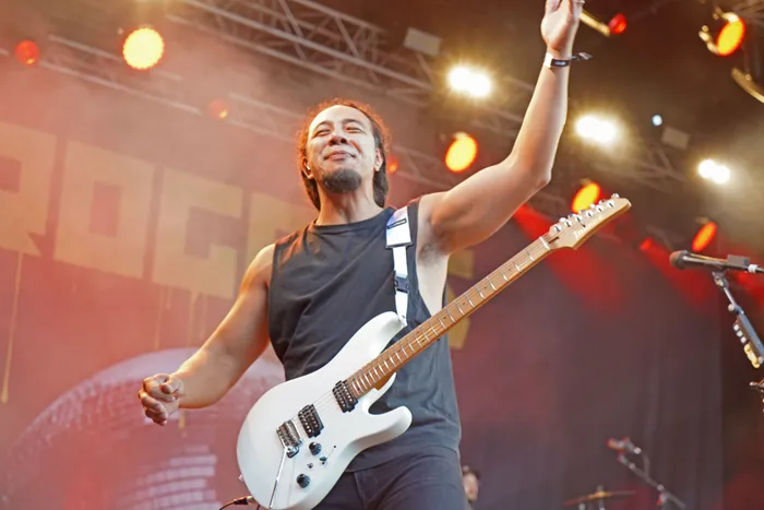 Rogers spielt mit einer weißen E-Gitarre auf einer hell erleuchteten Bühne und streckt einen Arm in die Luft.