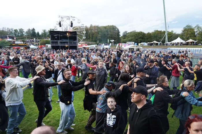 Eine Menschenmenge bei einem Musikfestival steht in Reihen mit den Händen auf den Schultern der Vorderleute vor einer Bühne und einem Riesenrad.
